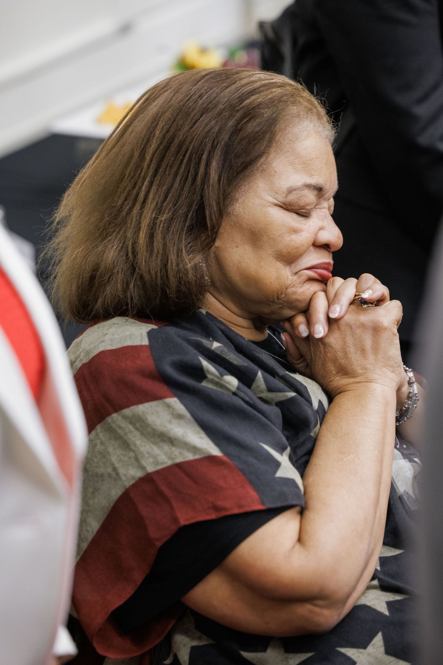 Elderly woman in American flag shawl praying with hands clasped and eyes closed at a patriotic event.