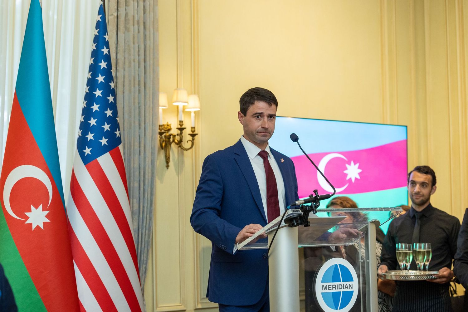Officials gather at a diplomatic meeting with US and Azerbaijan flags visible in an elegant conference room setting.