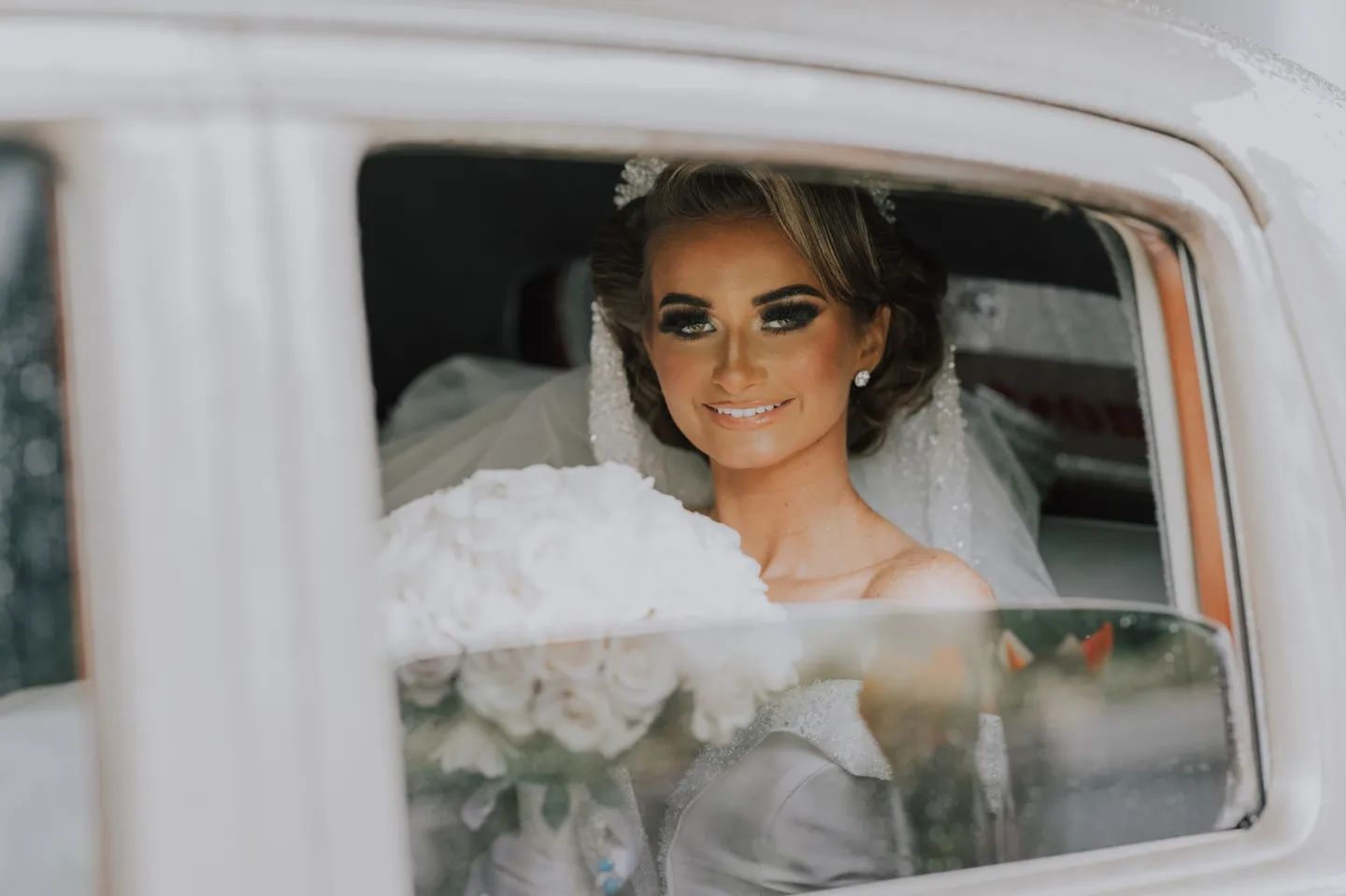 Bride sitting in vintage car smiling through window on wedding day.