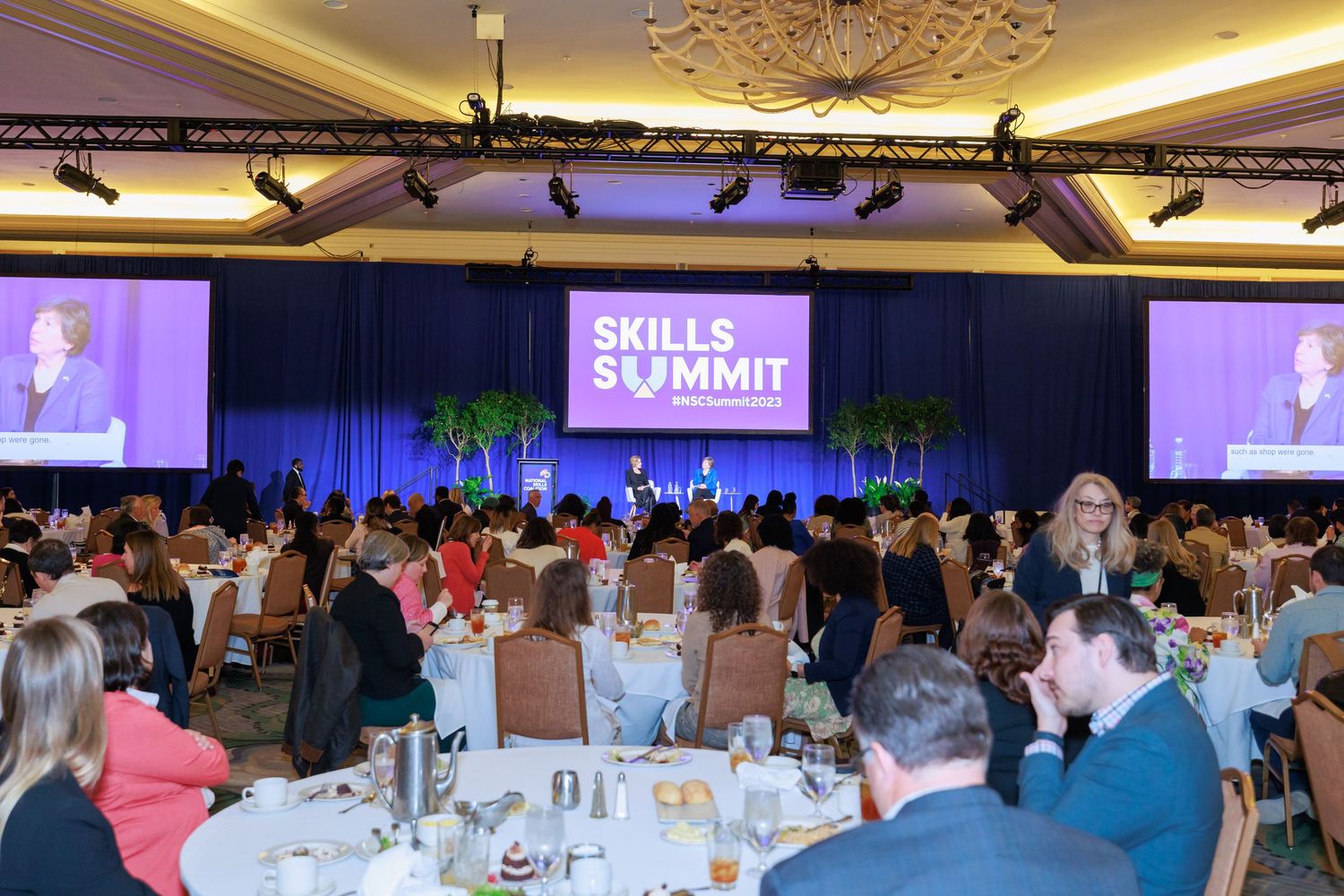 Large conference room filled with attendees during a corporate event with blue lighting and presentation screens.