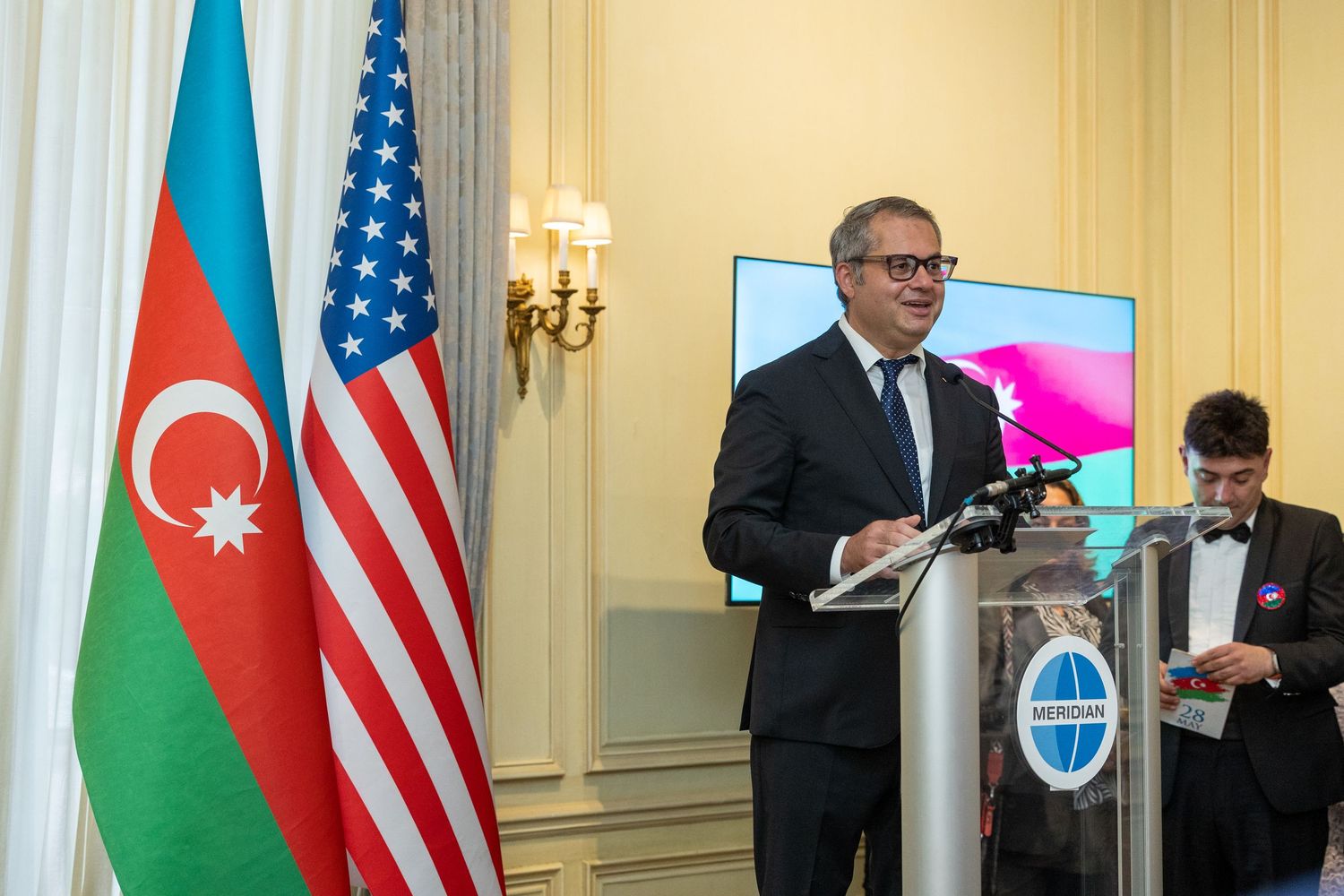 Two officials stand at podiums with US and Azerbaijan flags during a diplomatic meeting in an ornate room.