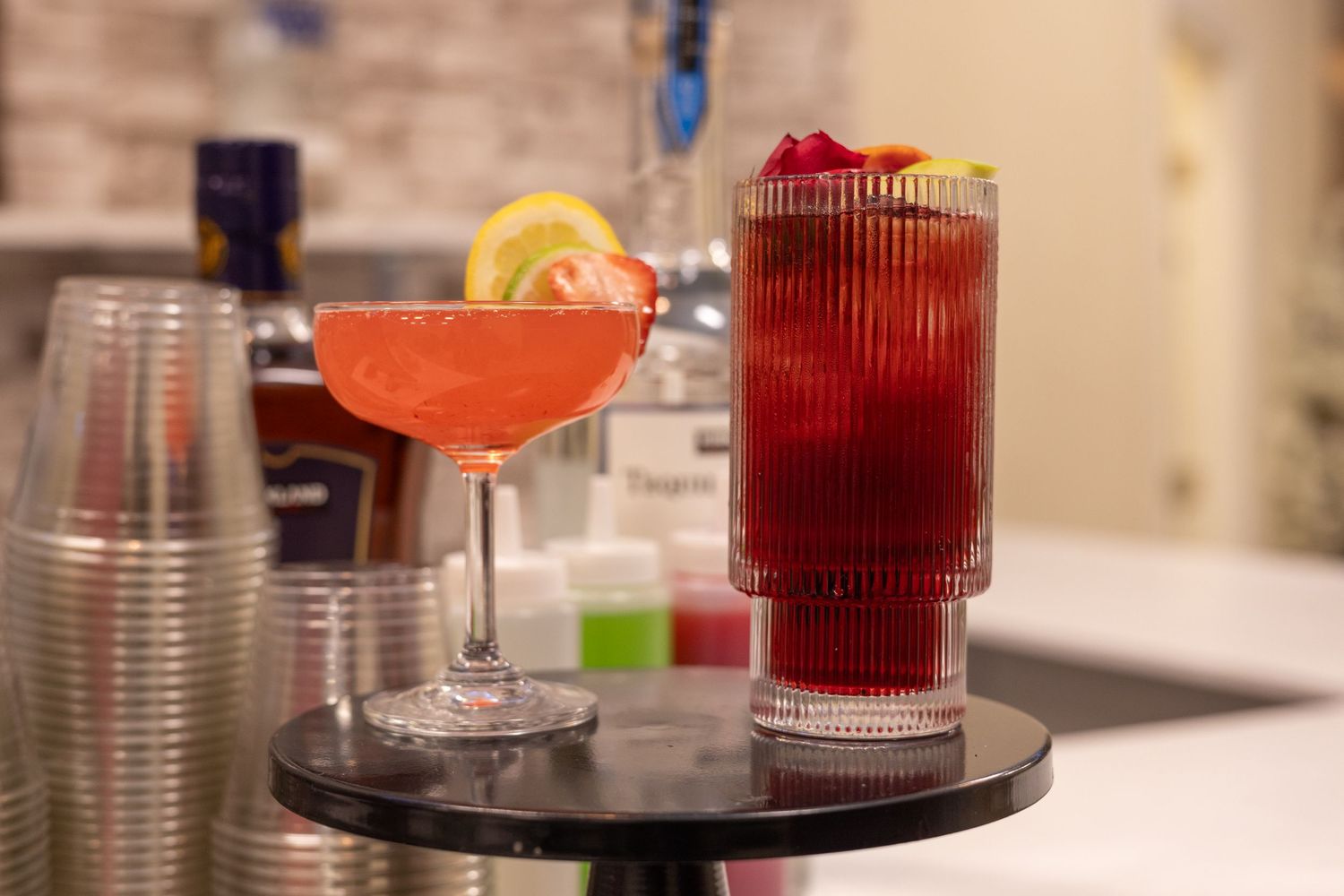 Two vibrant cocktails on a bar table, a orange margarita and a tall red drink garnished with cherries and lemon.