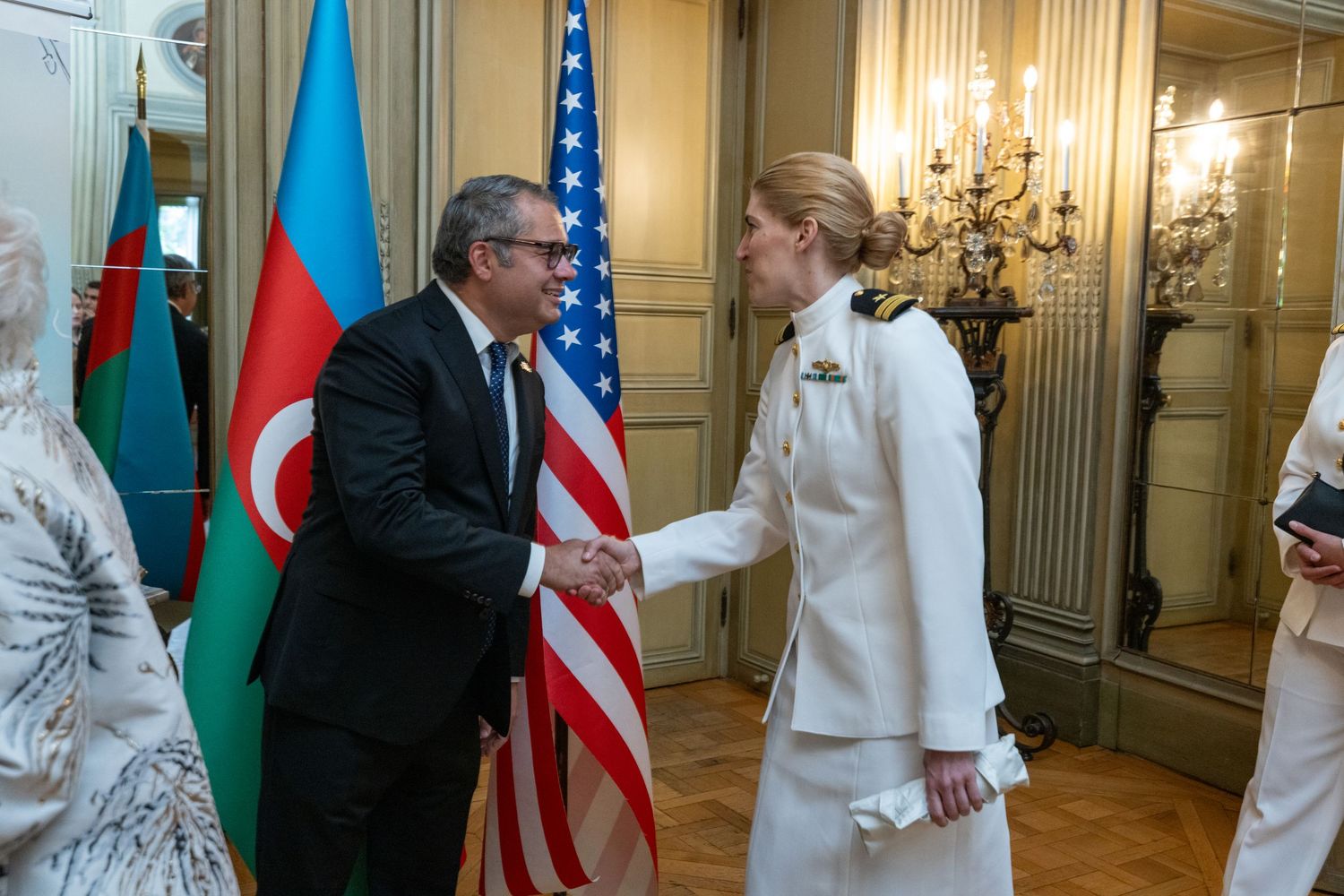 Diplomat in white suit shaking hands with official near Azerbaijan and US flags at formal reception.