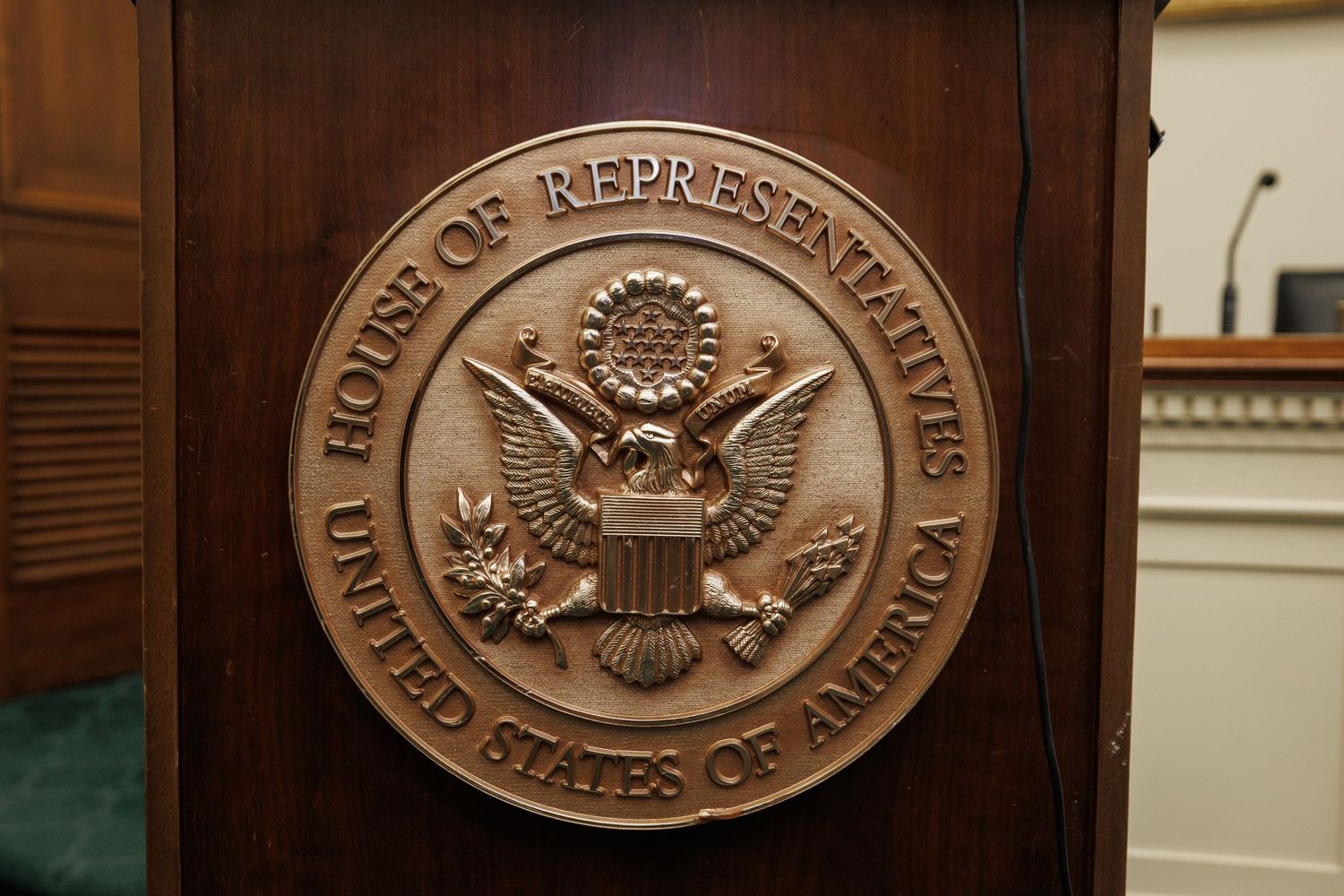 Bronze seal of the U.S. House of Representatives mounted on a wooden podium in a congressional hearing room.