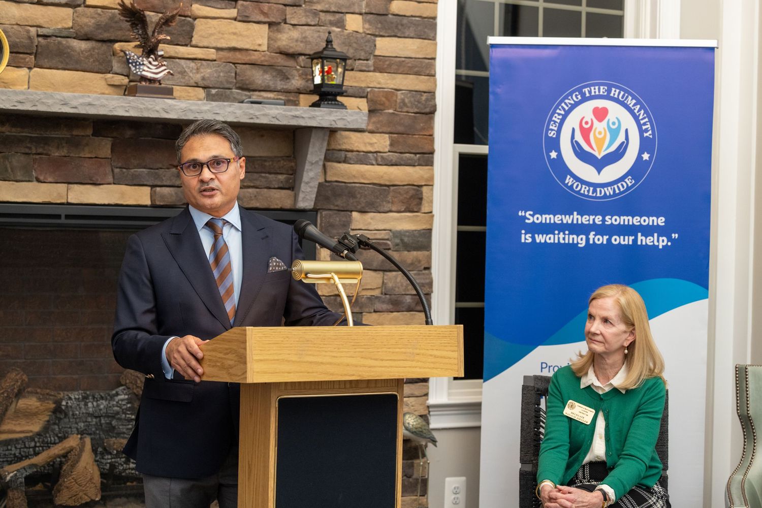 People stand at podiums during a presentation event with blue banners displaying government agency logos.