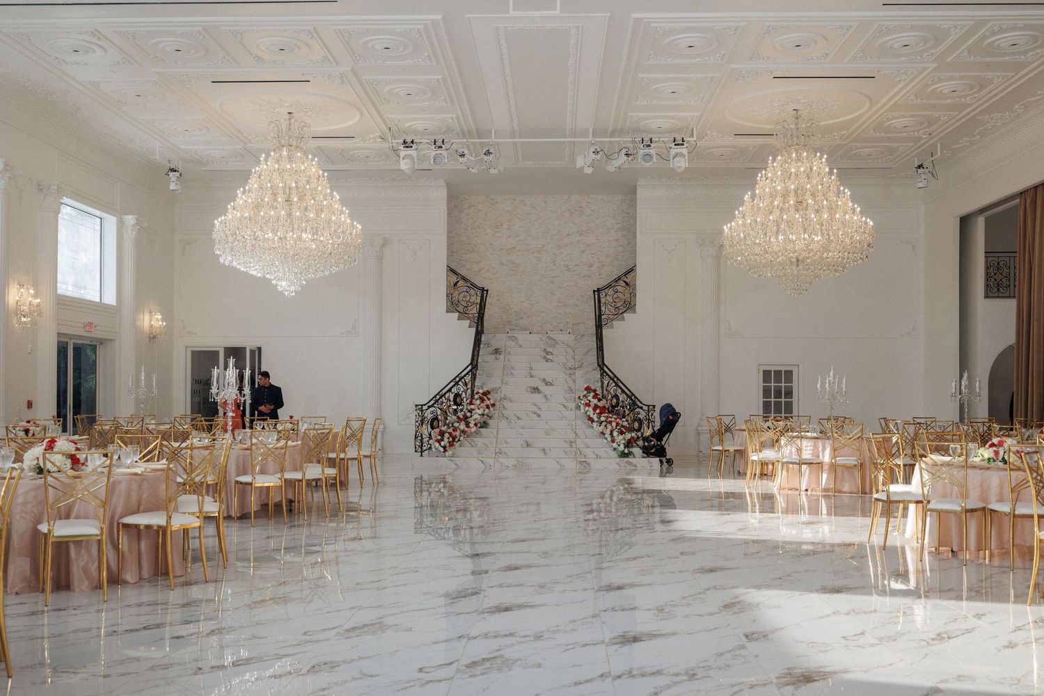 Elegant white ballroom with crystal chandeliers and round tables set for a formal event with floral centerpieces.