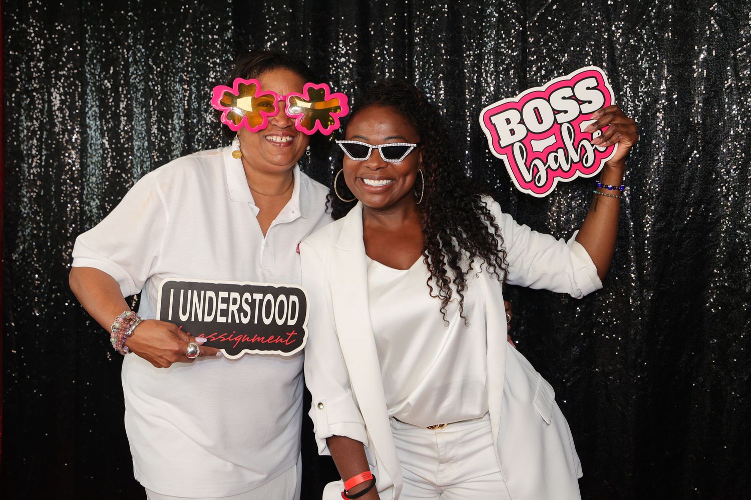 Two women in white outfits pose with photo booth props including Boss Babe sign against black sequin backdrop.