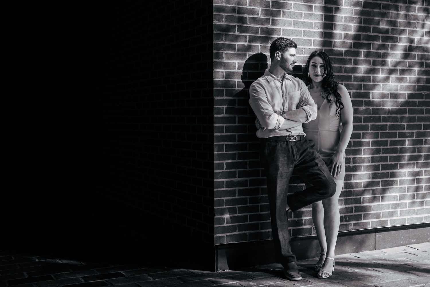Expecting couple stands in dramatic lighting against brick wall.