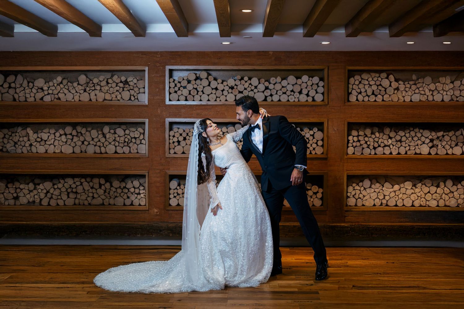 A couple shares a romantic moment in front of rustic wood-stacked wall in elegant wedding attire.