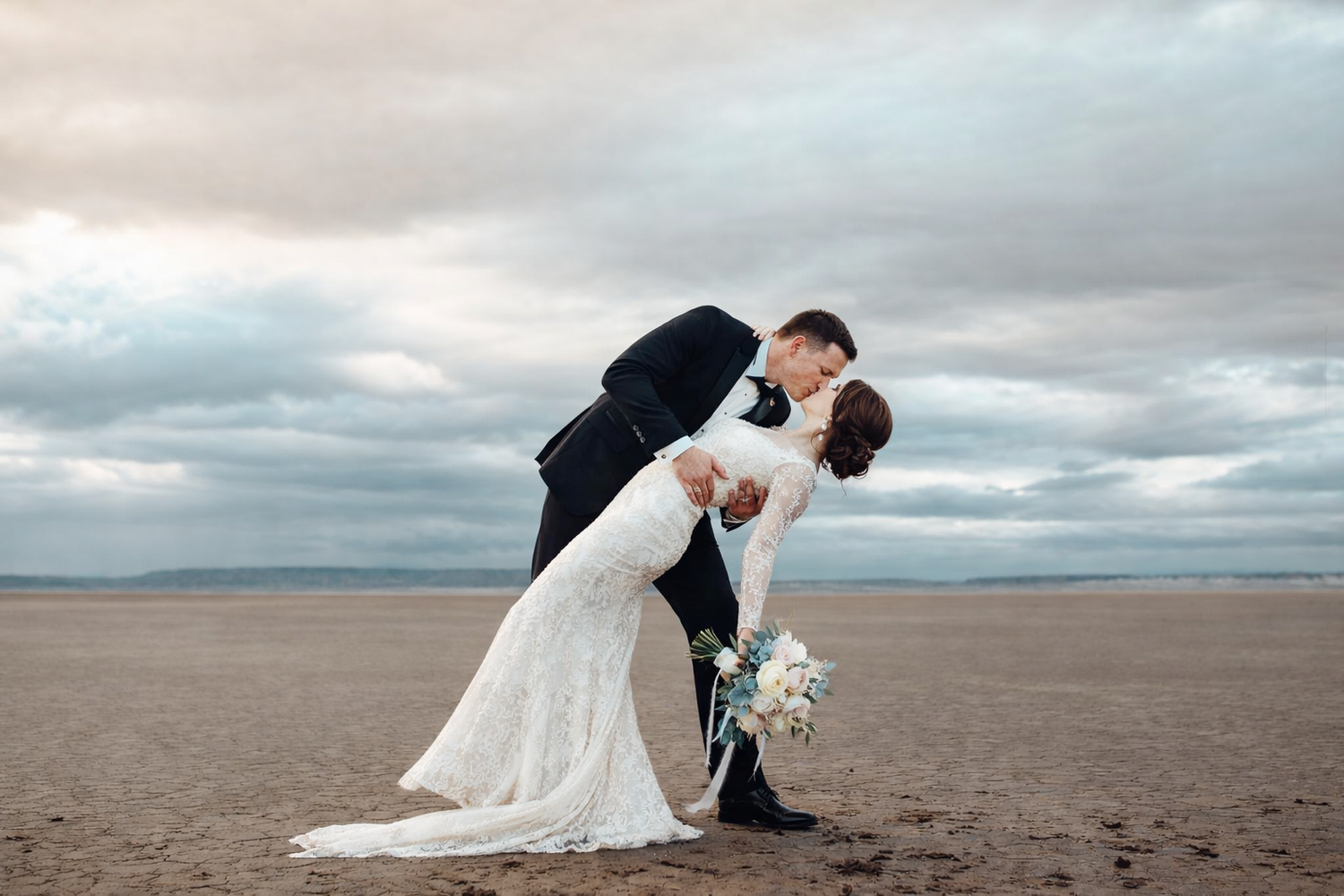 Romantic wedding couple dips and kisses on a dramatic cloudy beach at sunset.