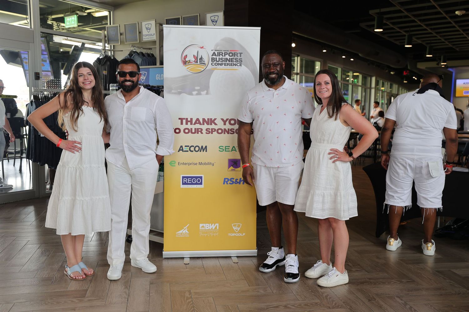 Four people in white attire pose together at an Airport Business event with sponsor banner backdrop.