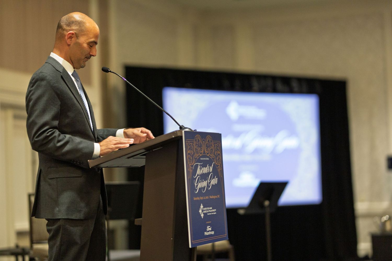 Person in suit standing at podium with presentation screen and chalkboard visible in background during business event.