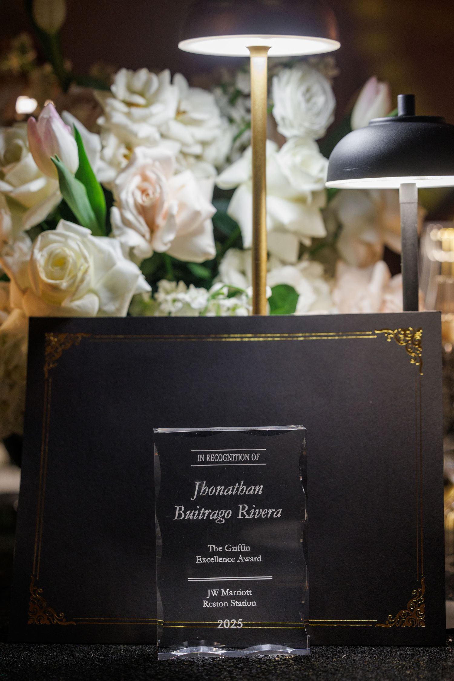 Black award plaques with engraved names displayed on a table with white floral decorations and lamps in the background.