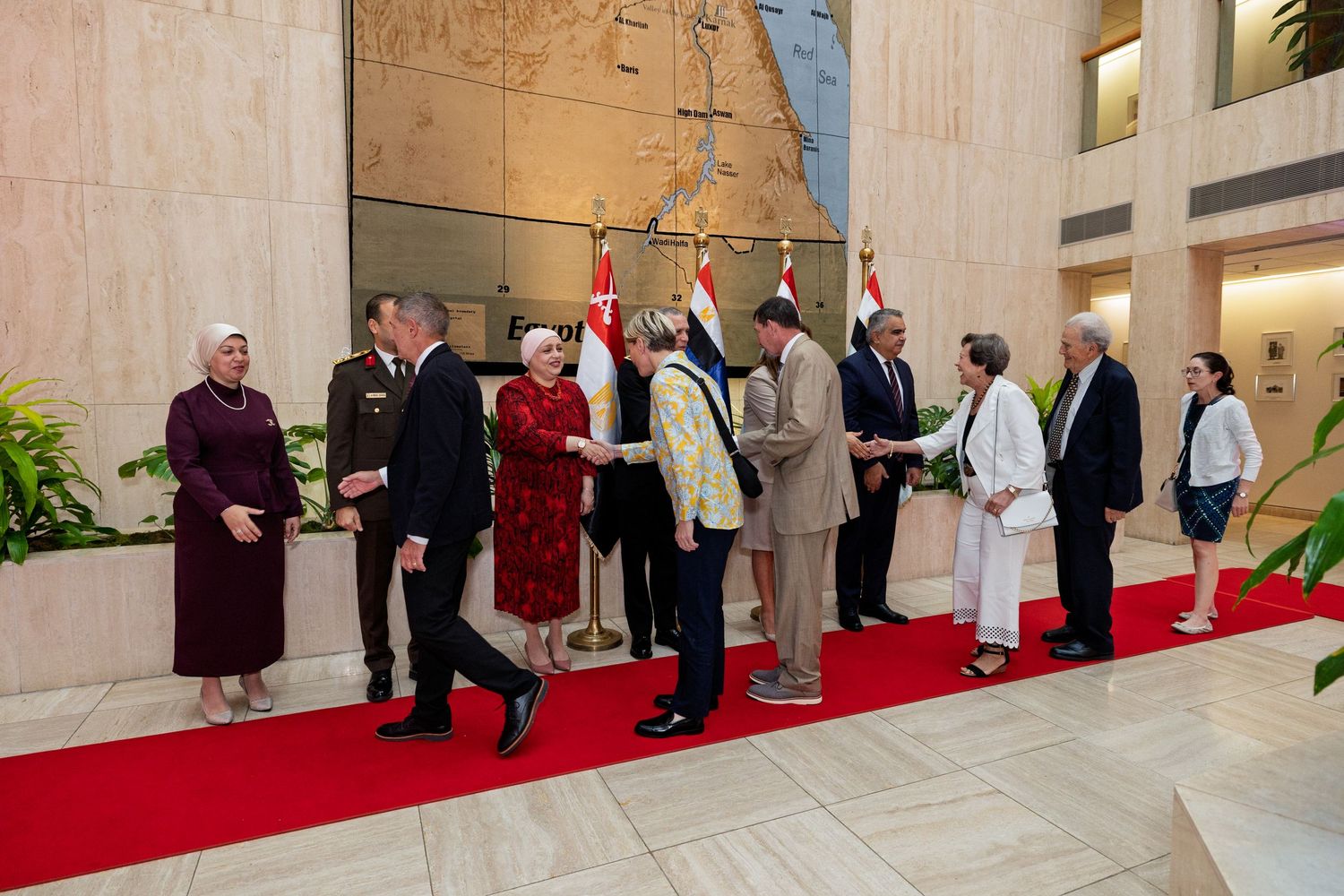 A row of people in formal attire stand along a red carpet in an ornate indoor venue during a ceremonial event.