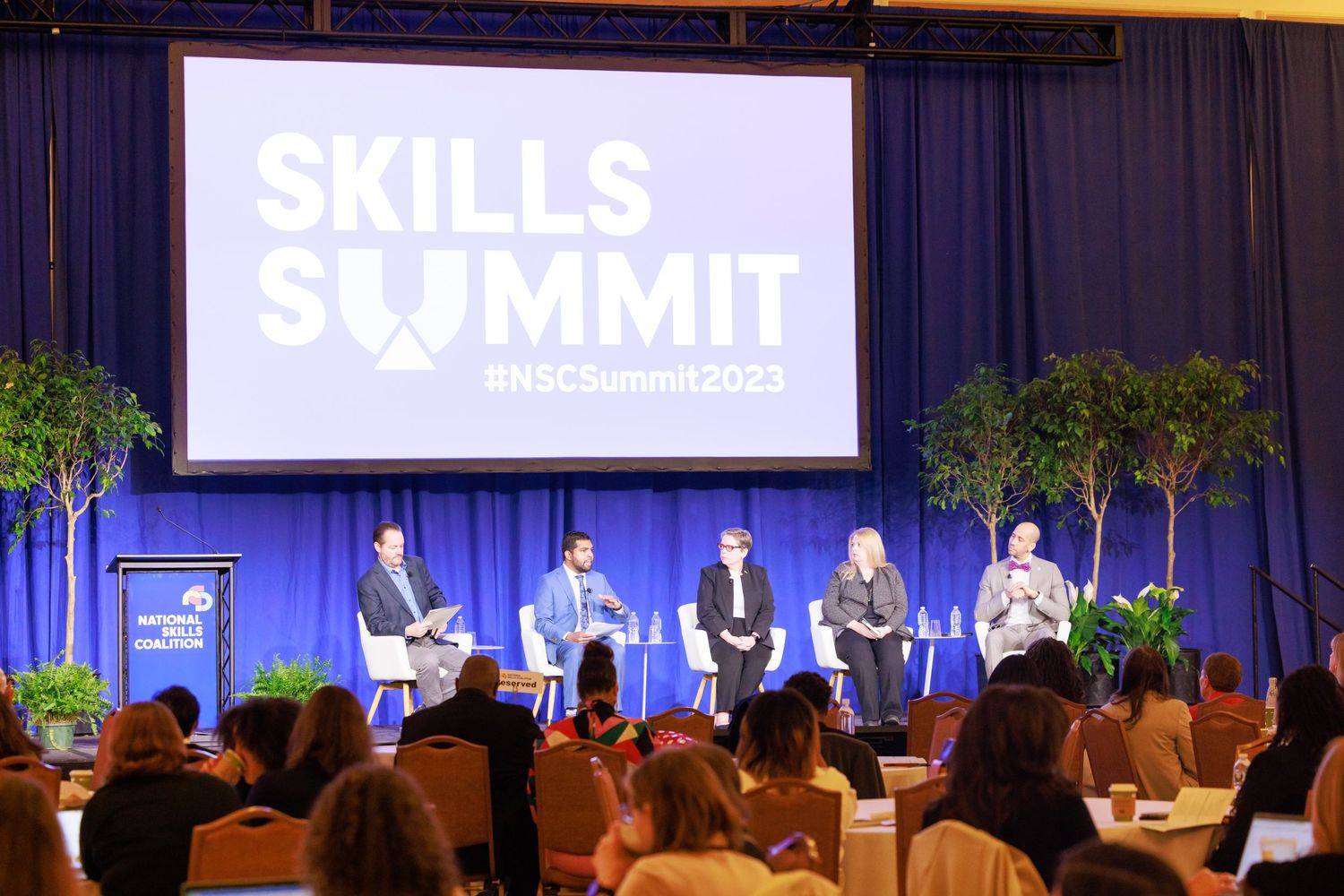 Business conference panel discussion with speakers seated at tables on stage with blue backdrop and Skills Summit signage.