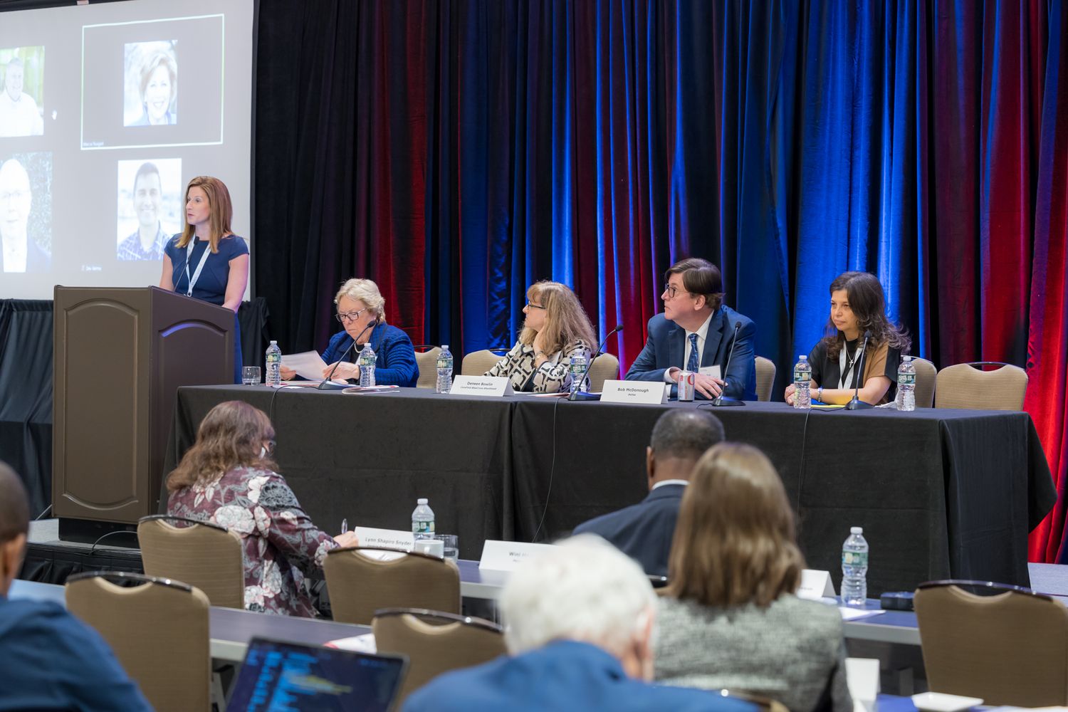 People seated at conference tables with red and blue backdrop during a business meeting.