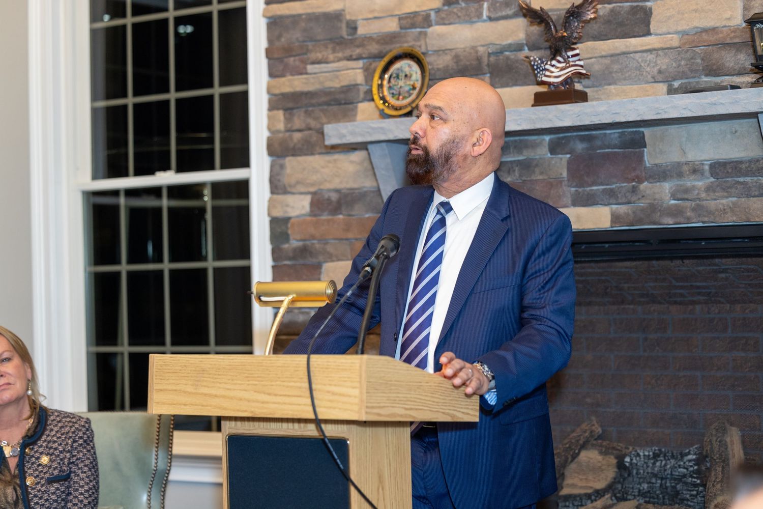 A speaker in a blue striped tie addresses an audience from a wooden podium in front of a stone fireplace.