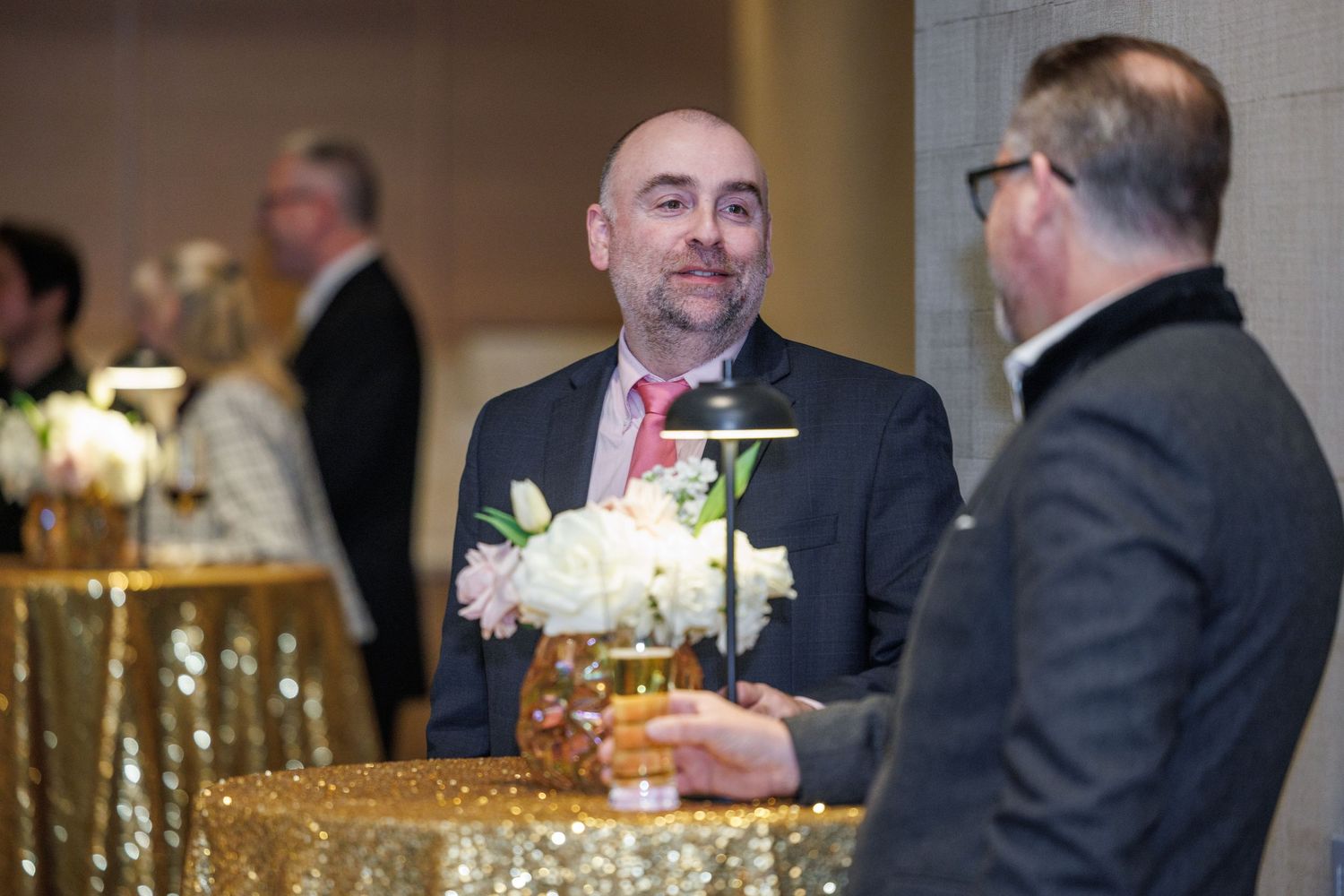 Two people in formal attire having a conversation at an event with a gold sequined tablecloth and floral centerpiece.