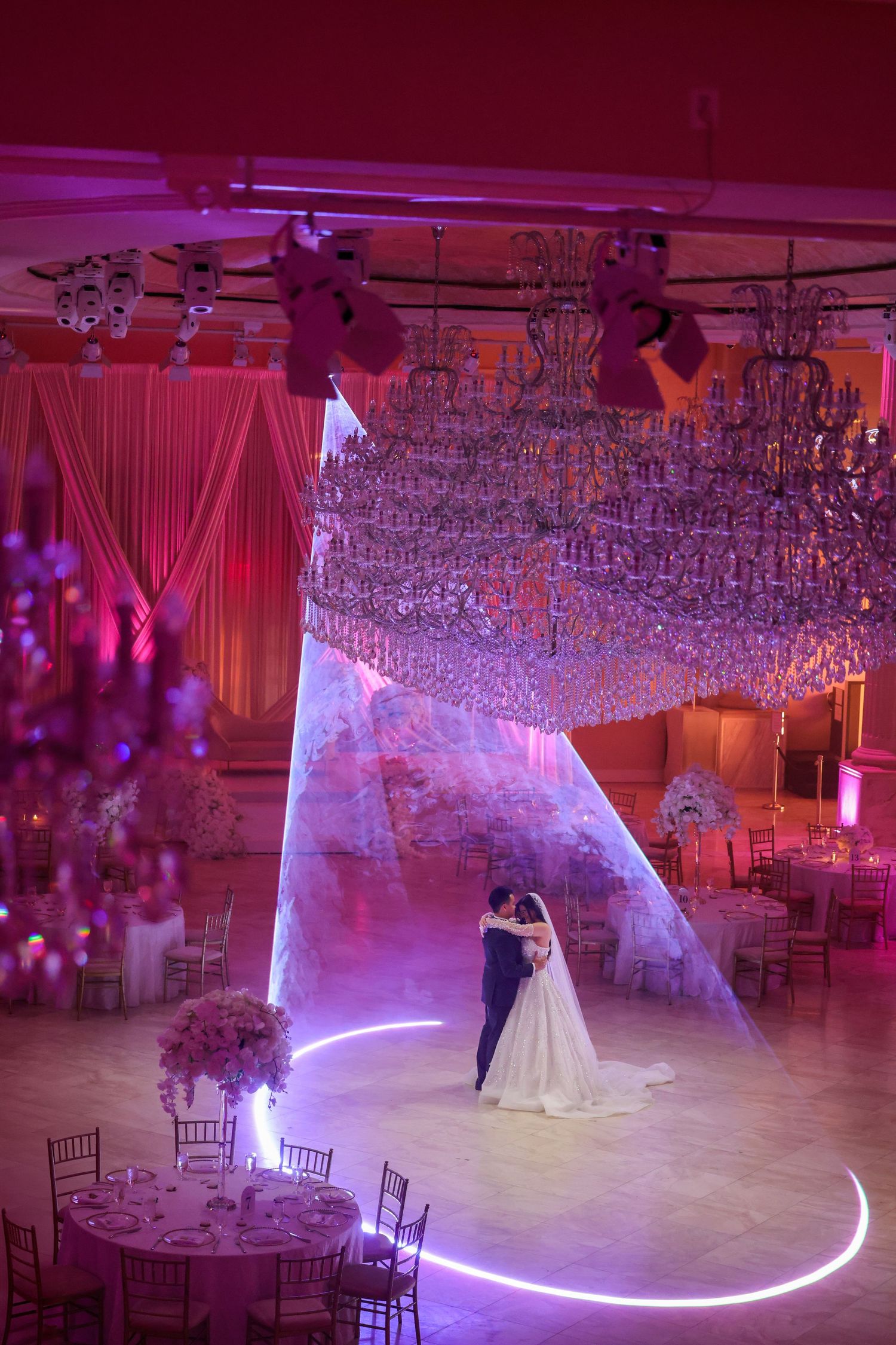 Elegant wedding reception ballroom decorated with crystal chandeliers, white drapes, and illuminated curved LED floor lighting.