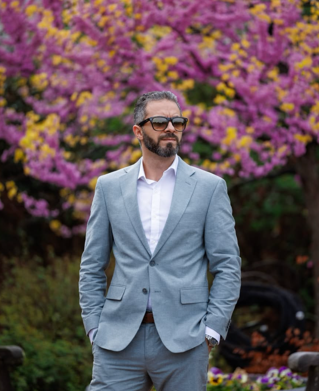 Man in light gray suit and sunglasses standing confidently before vibrant pink and yellow flowering trees.