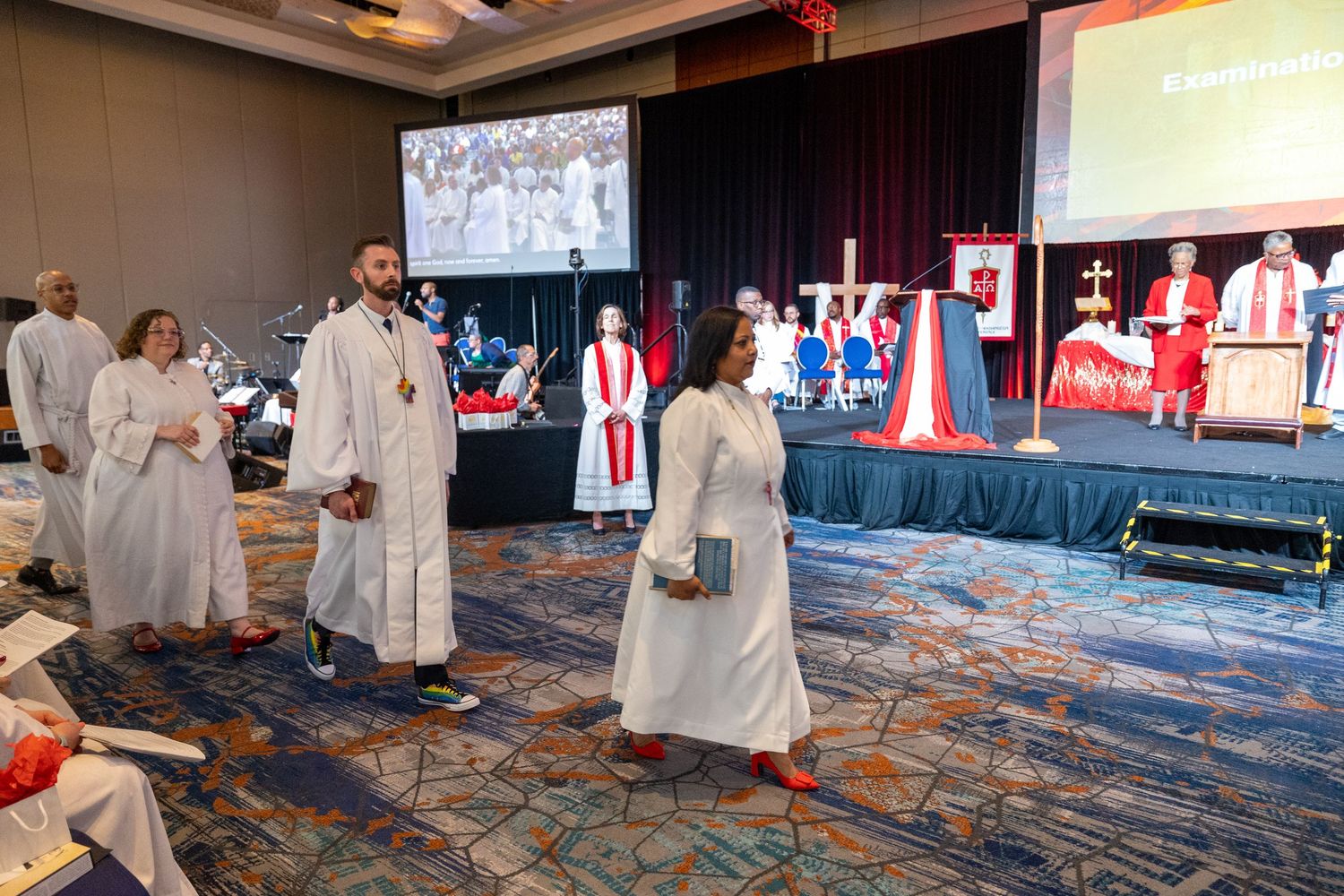 Religious ceremony participants in white robes gather on a carpeted floor in front of a stage with red and blue decorations.