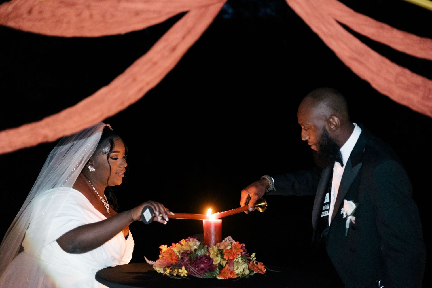 A couple lights a unity candle during their wedding ceremony beneath coral-colored fabric draping.