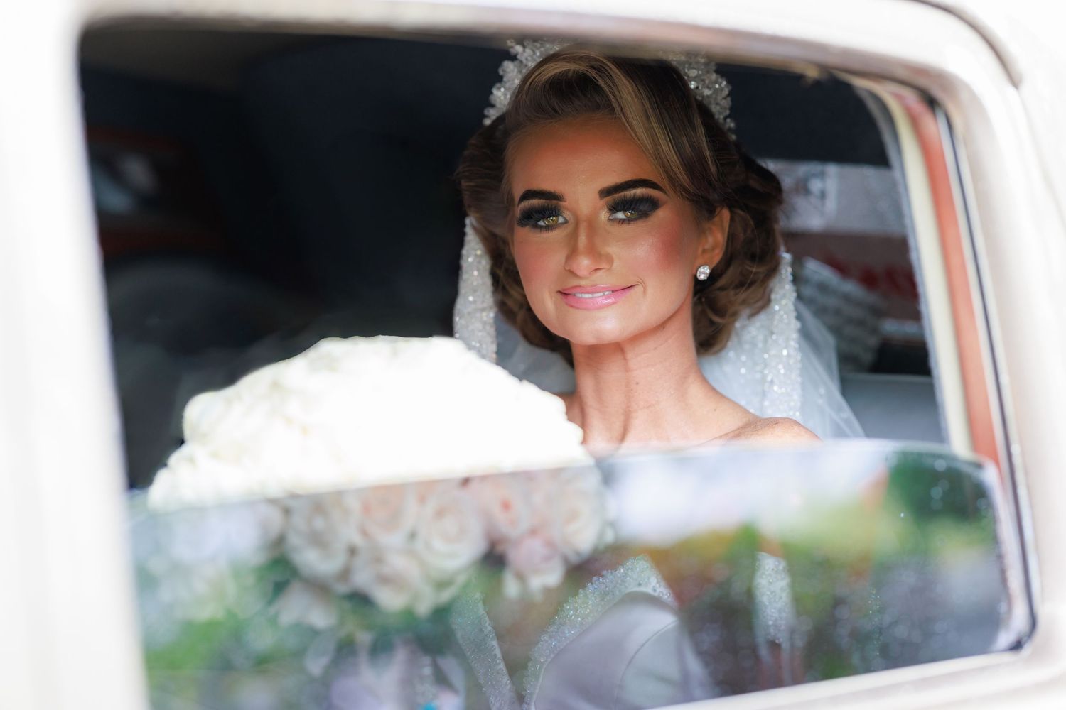 A bride and bridesmaid smile while seated in a vintage car decorated with white flowers and ribbons.