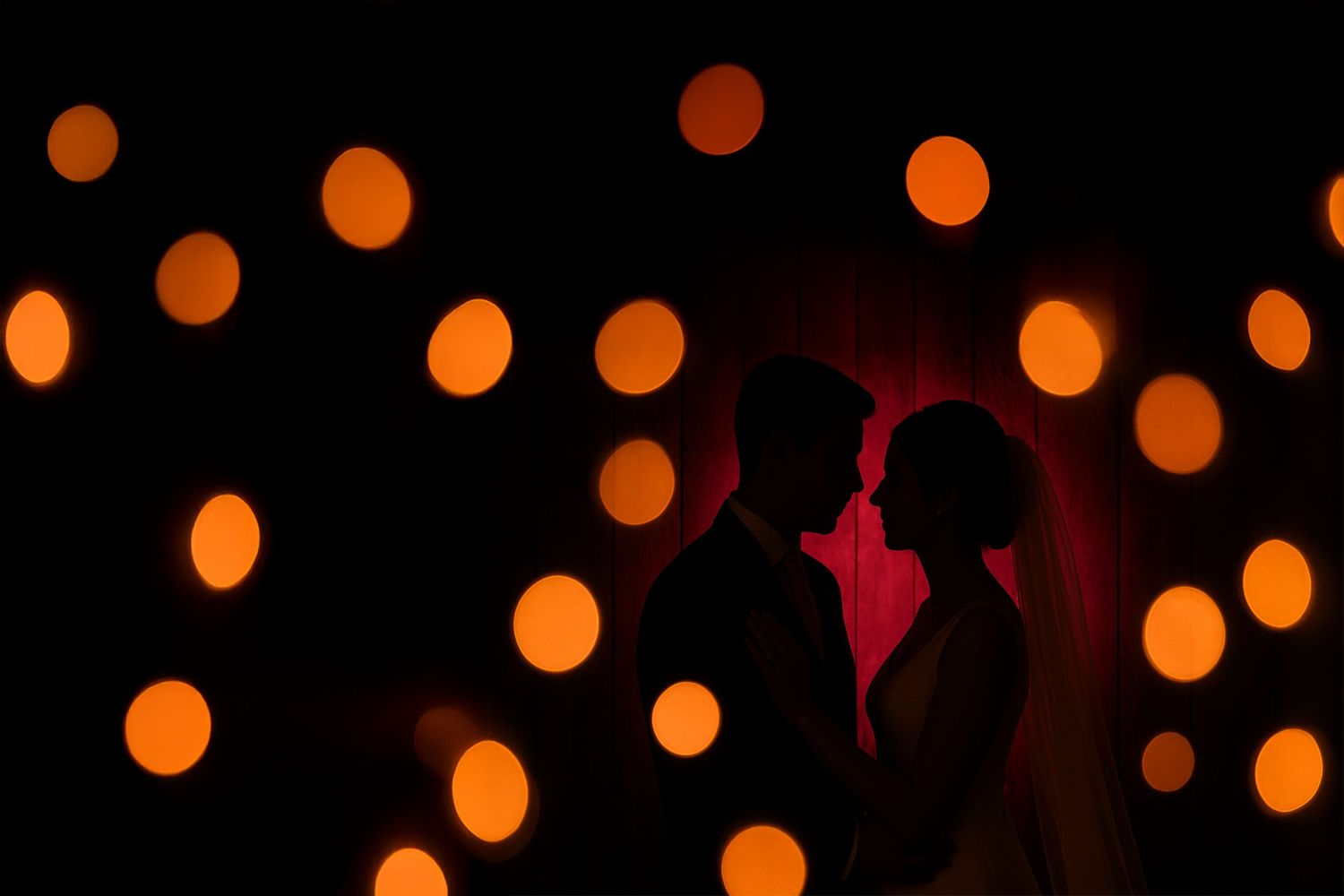 Silhouettes of a couple stand against a backdrop of glowing orange bokeh lights creating a romantic atmosphere.