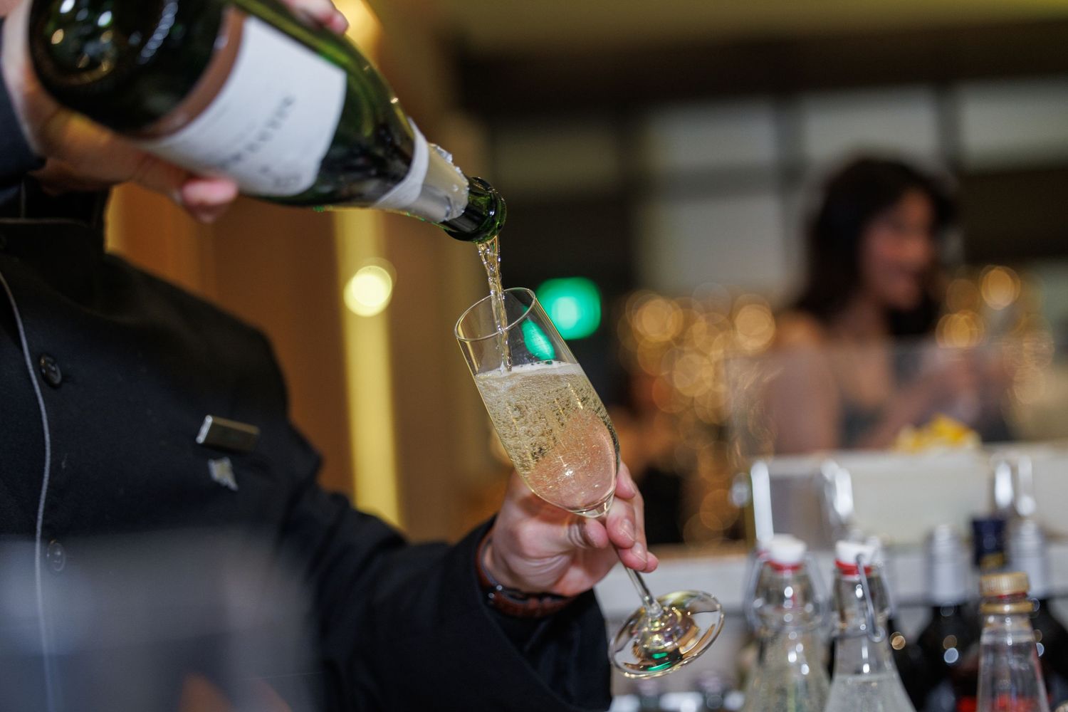 Bartender pouring champagne into a flute glass at an elegant event with bottles and guests in background.