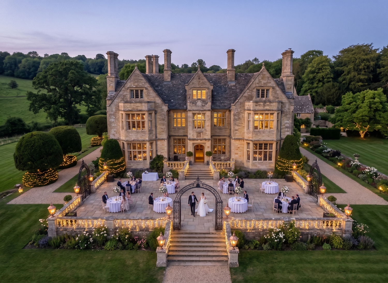 Aerial twilight view of an elegant stone manor house with wedding reception on illuminated terrace surrounded by manicured gardens.
