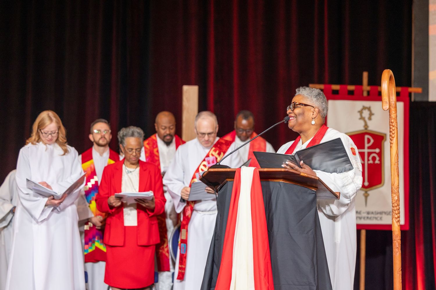 Performers in red and white robes stand in line on stage during a theatrical production.