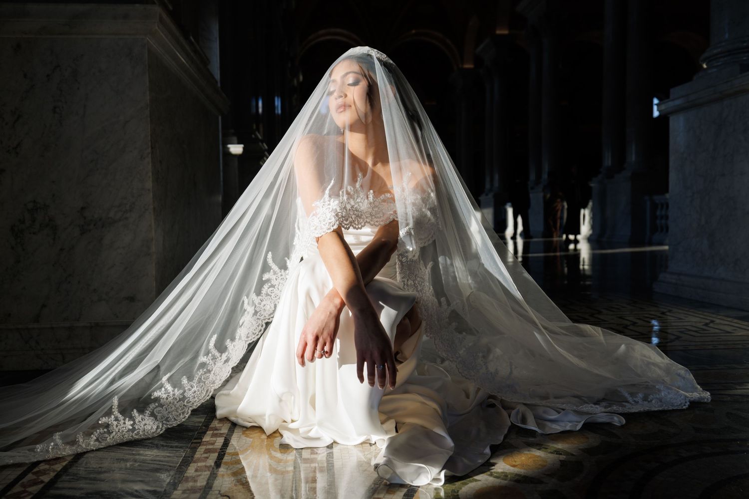 A flowing white wedding dress train dramatically spreads across a dark floor in artistic bridal photography.