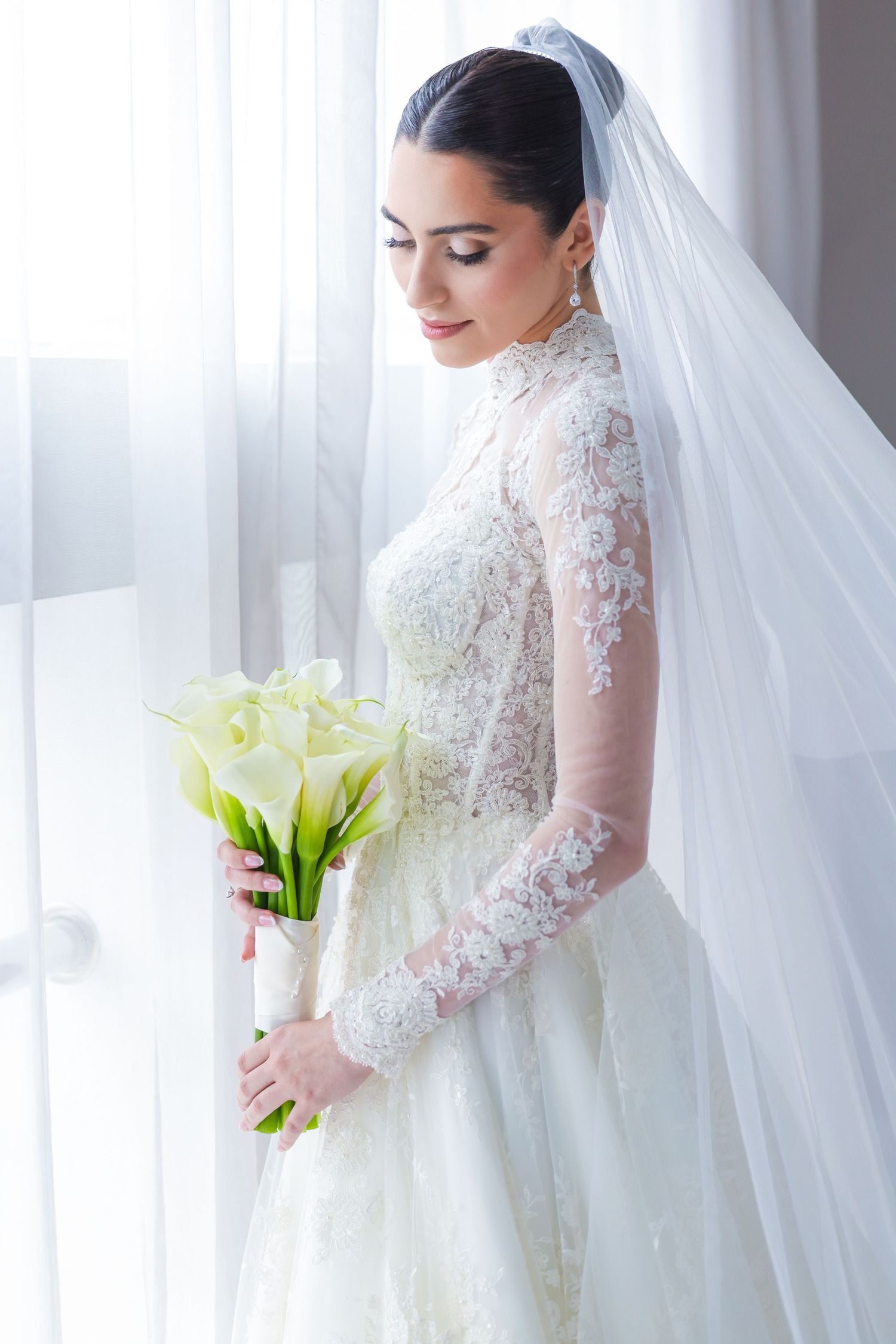 Elegant bride in long sleeve lace gown holding calla lily bouquet by window.
