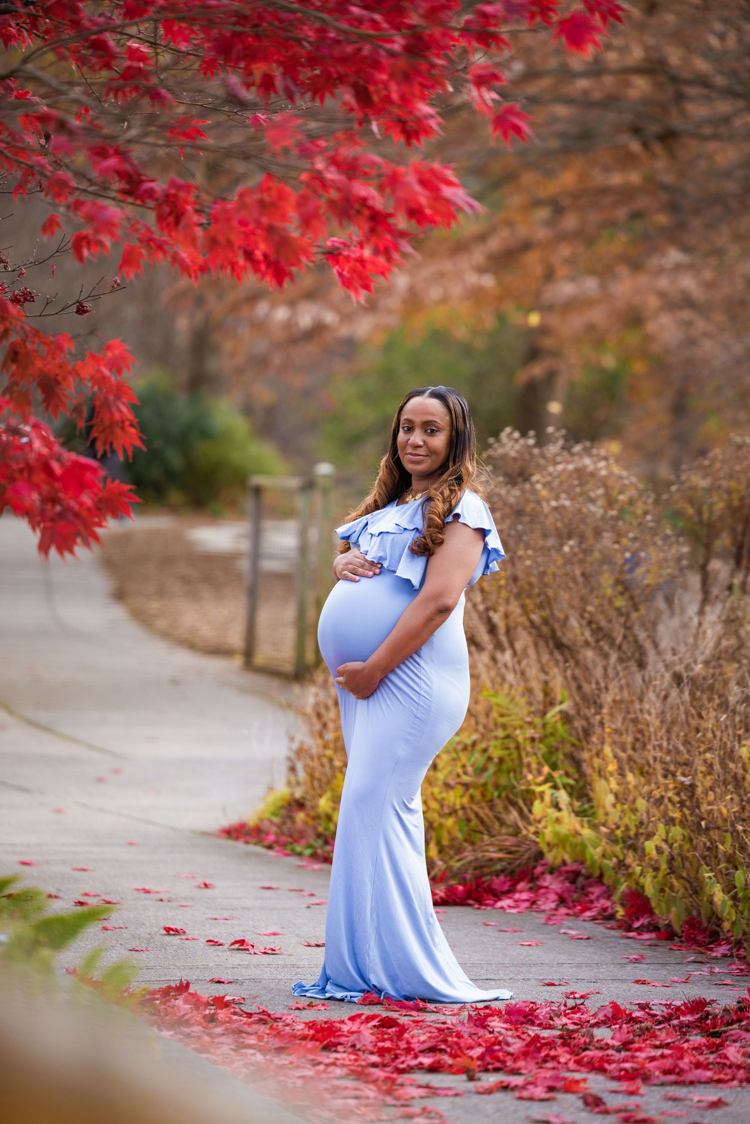 Maternity portrait with red autumn leaves framing woman in flowing blue dress.