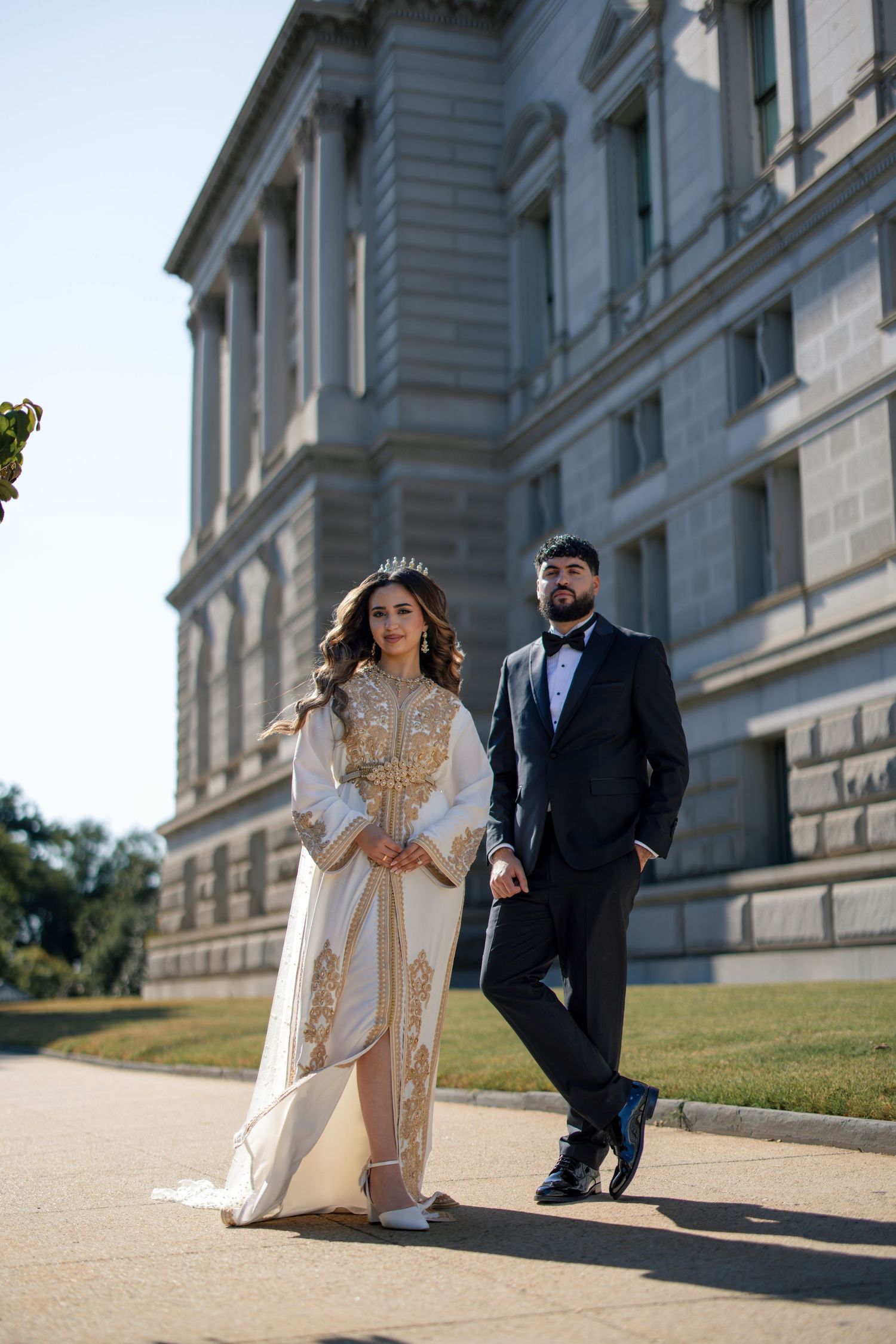 Elegantly dressed couple poses outside grand historic building with columns and stone architecture on sunny day.
