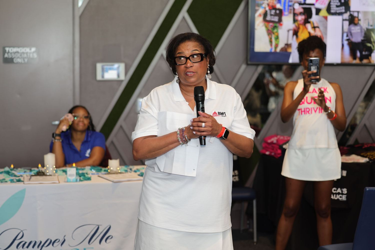 Woman in white outfit speaks into microphone at indoor event with attendees and colorful backdrop.