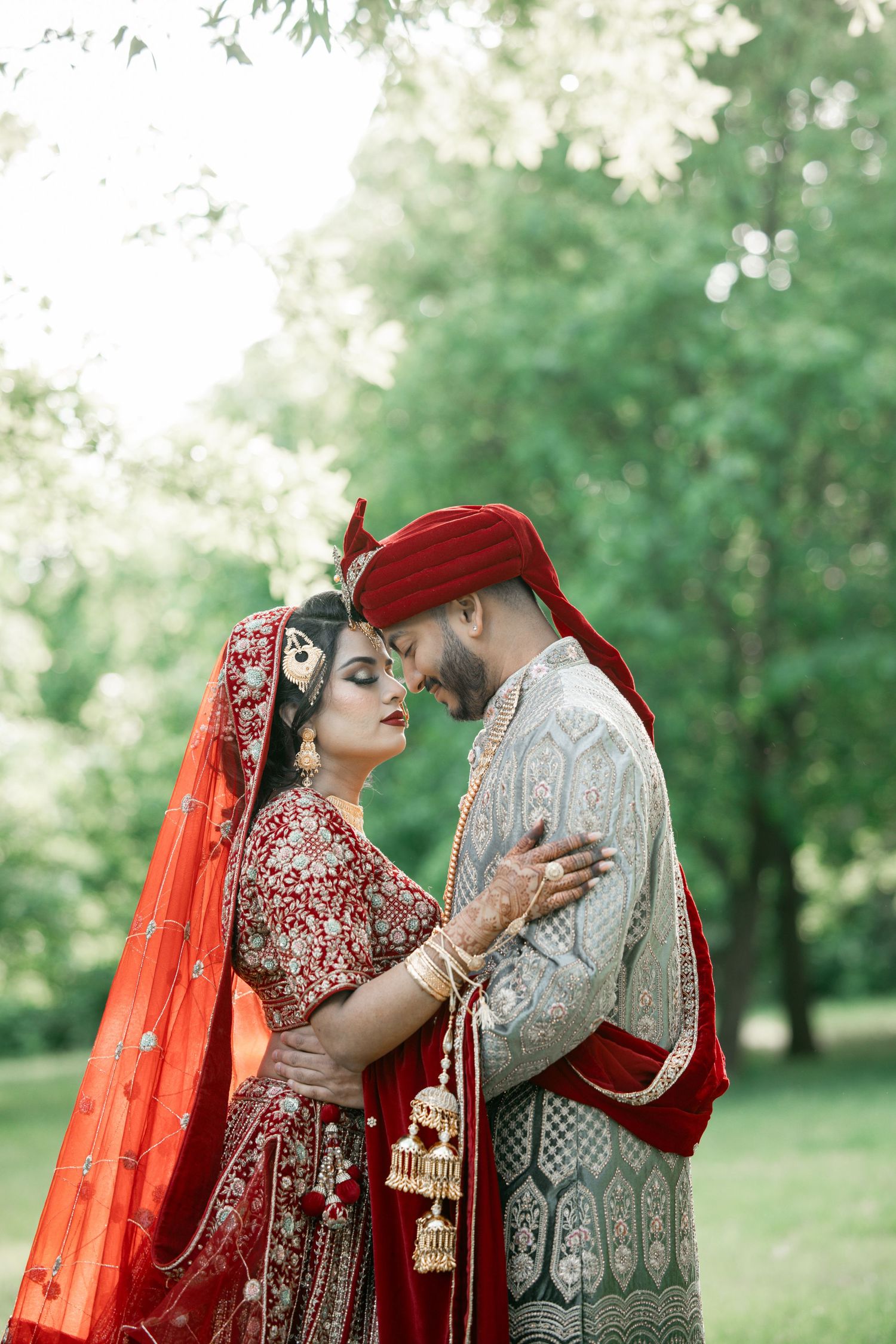 Traditional South Asian wedding couple in ornate red and gold attire embracing outdoors.