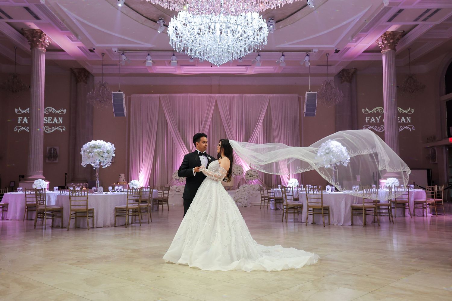 Elegant ballroom setup with crystal chandeliers and pink uplighting for a wedding reception.