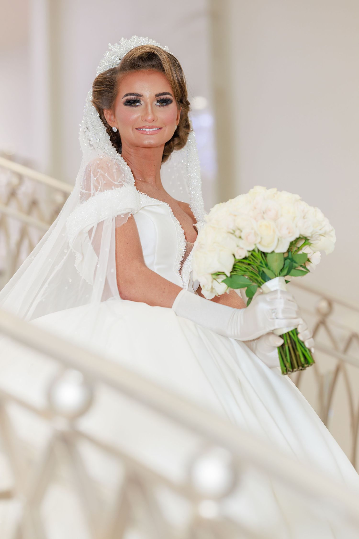 Elegant bride in white lace gown holds stunning white bouquet while posing on grand staircase.
