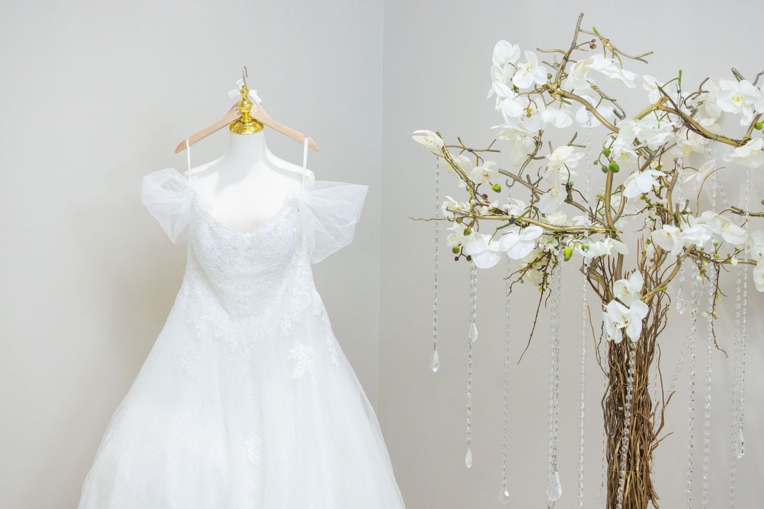 White christening gown hangs elegantly beside delicate white floral arrangement in soft natural light.