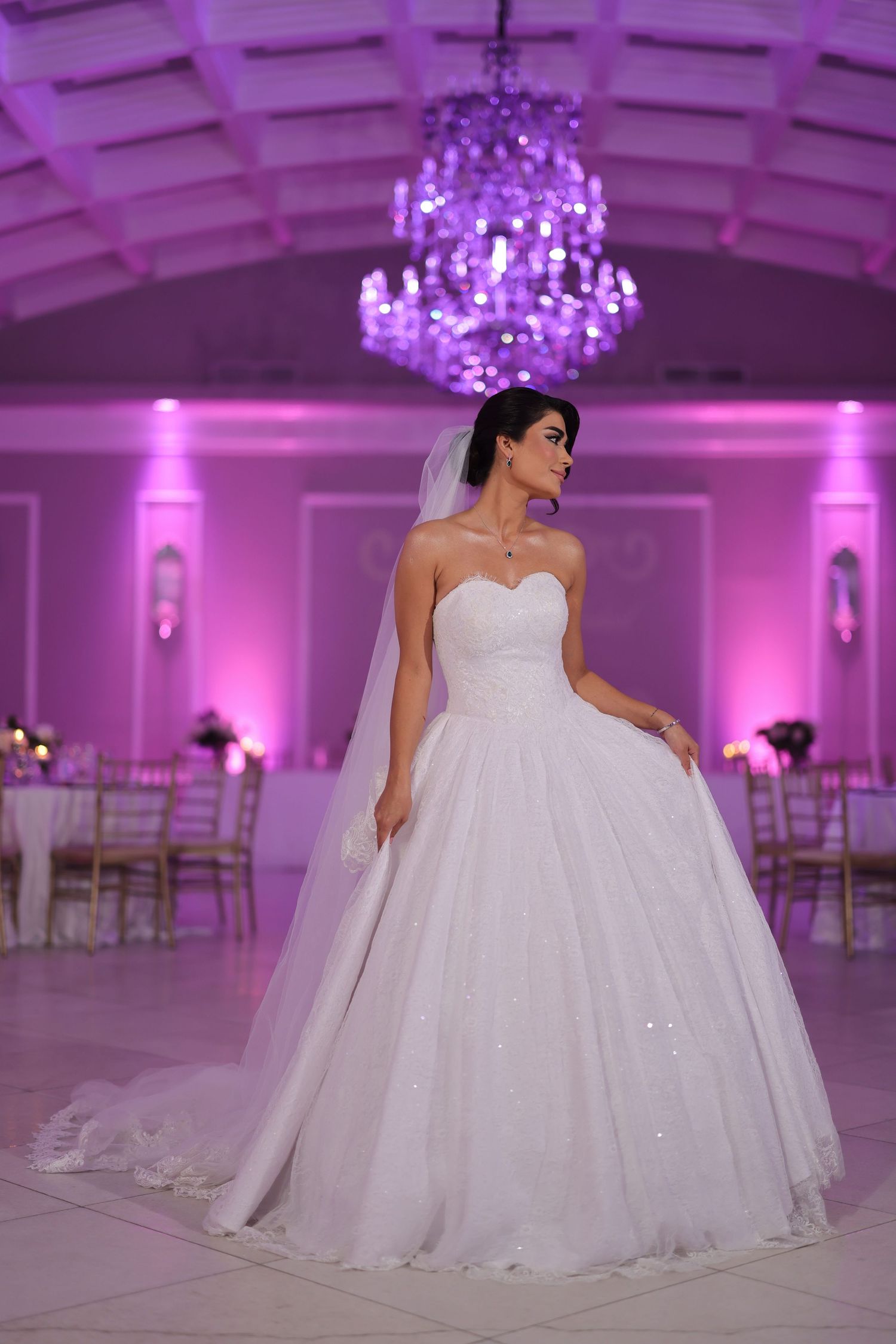 Elegant white ballgown wedding dress in purple-lit ballroom with crystal chandeliers.