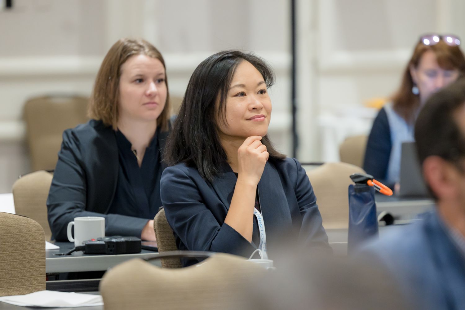 Attentive professionals participate in a business conference session while seated at tables in a modern meeting room.
