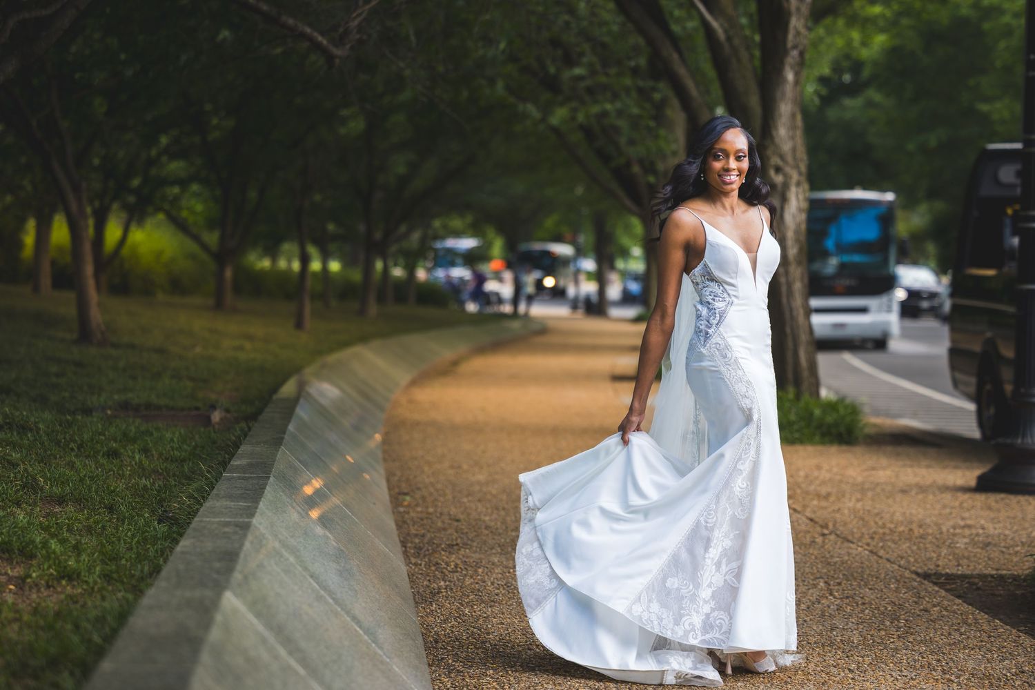 Someone in a flowing white dress walks along a tree-lined path near parked cars in a scenic outdoor setting.