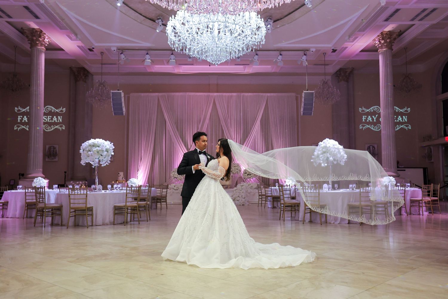 Elegant ballroom with crystal chandeliers and pink uplighting features white tables and chairs set for a formal event.