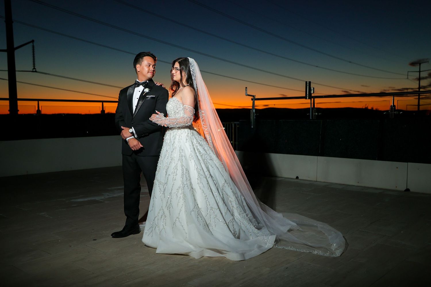 Silhouetted couple poses against a dramatic orange and purple sunset sky with power lines in the background.