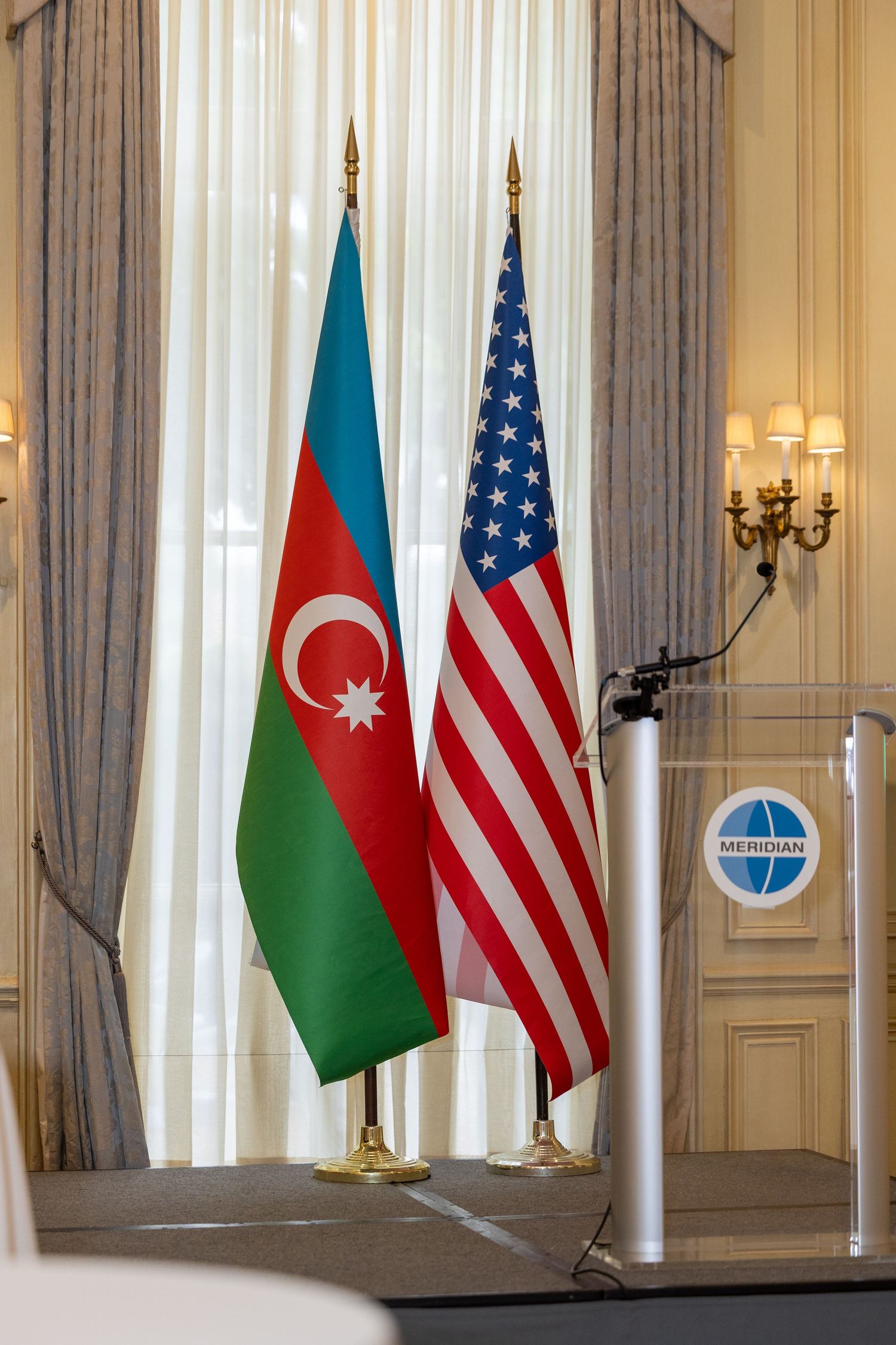 American and Azerbaijani flags stand together on display in an elegant room with cream curtains and gold accents.