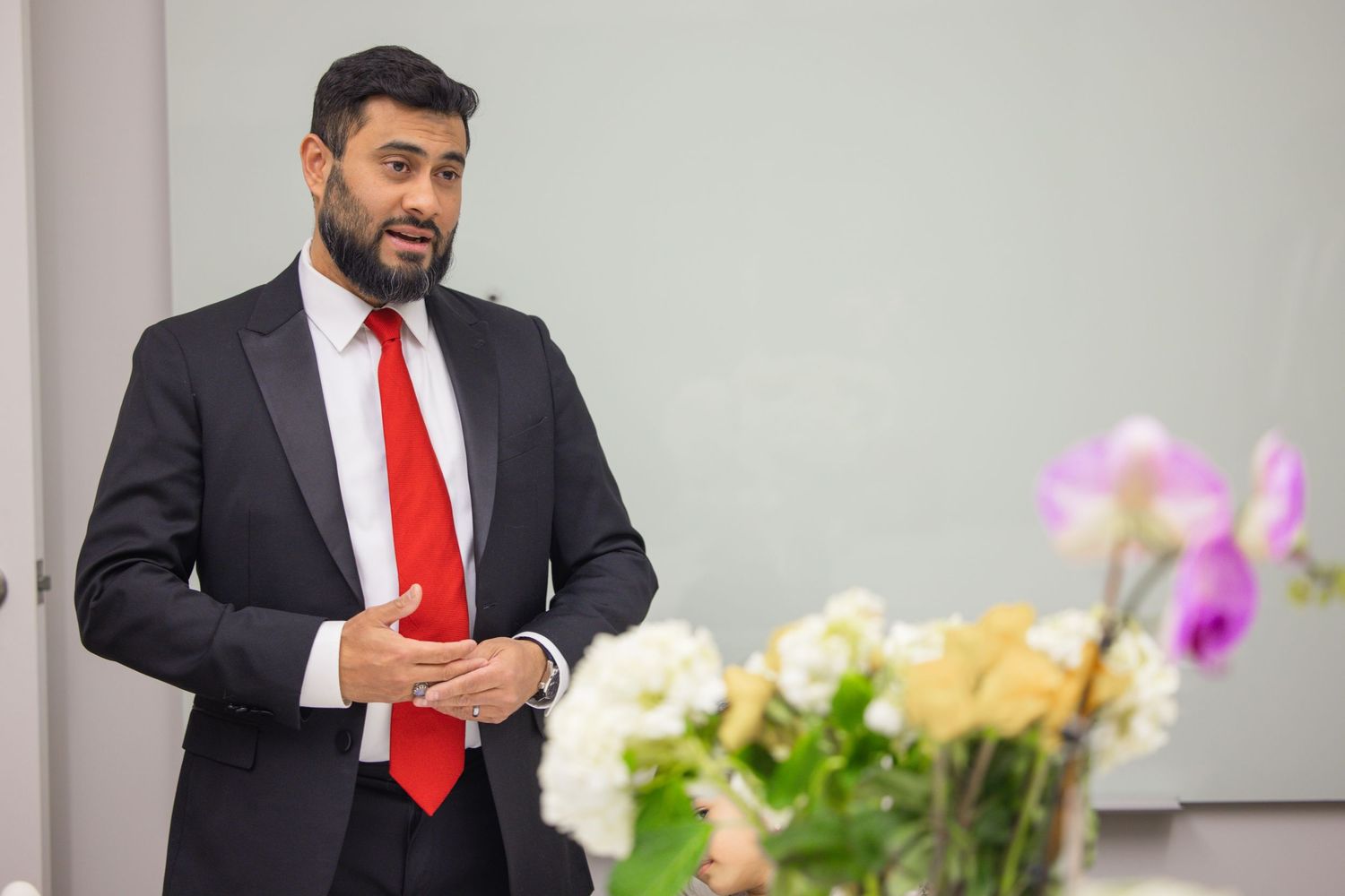 Professional in a dark suit and red tie stands in an office setting with flowers and decor in the background.