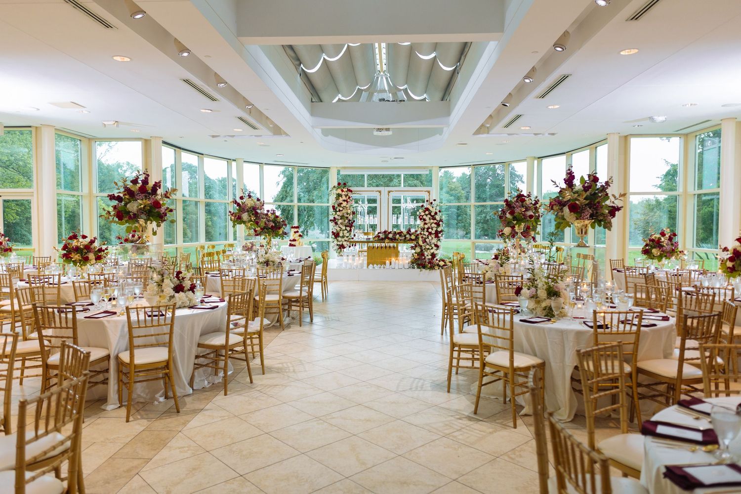 Elegant wedding reception venue with glass conservatory walls, wooden chairs and floral centerpieces arranged in rows.