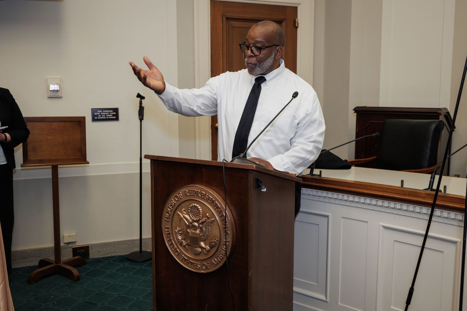 Man in white shirt and tie speaks at ornate wooden podium with official seal in formal government chamber.