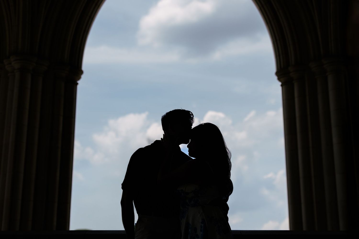 Silhouettes of two people standing in a stone archway against a bright cloudy sky.